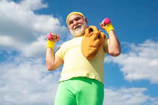 Cura del corpo e assistenza sanitaria. Uomo anziano che pratica sport su sfondo cielo blu. Felice uomo anziano con i manubri che guarda la macchina fotografica. Vecchio uomo maturo esercizio con manubrio . — Foto Stock