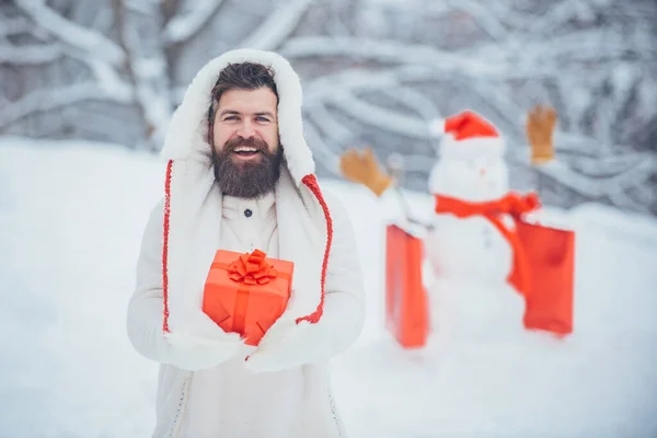 La preparazione di Natale - l'uomo barbuto divertente con scatola di regalo rossa fa il pupazzo di neve. Godetevi la natura invernale. Holly jolly swag Natale e noel . — Foto Stock