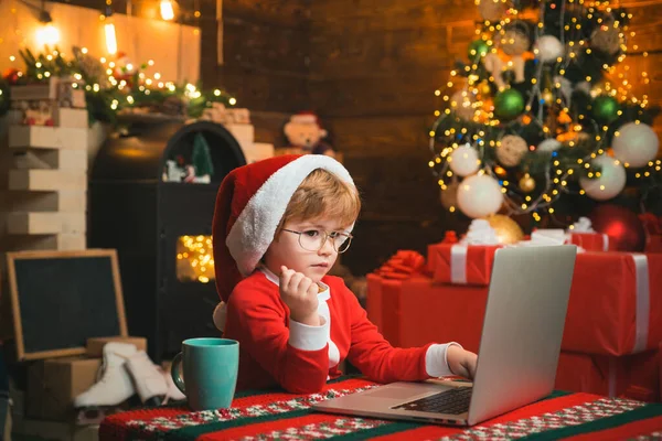 İnternetten çocuklar için Noel alışverişi. Noel çocuğu. Mutlu küçük gülümseyen çocuk Noel Baba kıyafetleri giyiyor ve dizüstü bilgisayarına mektup yazıyor.. — Stok fotoğraf