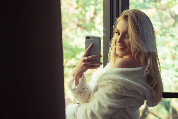 Ráno krásná mladá žena s telefonem u okna. Chladná žena v pyžamu, směje se doma. — Stock fotografie