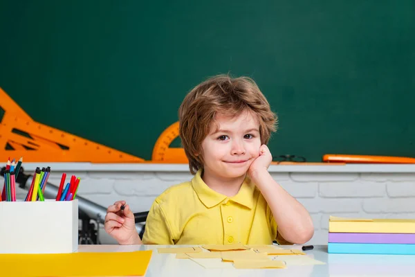 Wracać do szkoły. Szczęśliwy uśmiechnięty uczeń rysujący przy biurku. Przyjazne dziecko w klasie przy biurku z tablicą. Dzieciak uczy się w klasie na tle tablicy.. — Zdjęcie stockowe