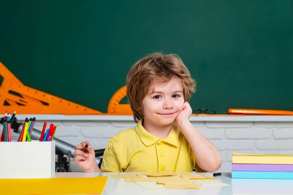 Terug naar school. Vrolijk lachende pupil tekening aan het bureau. Vriendelijk kind in de klas vlakbij schoolbord bureau. Kind leert in de klas op de achtergrond van schoolbord. — Stockfoto