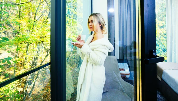 Woman with morning coffee. Portrait of relaxed girl looking outside through a window and holding a coffee in the living room at home. — Stock Photo, Image