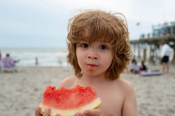 Retrato engraçado de um menino ruivo incrivelmente bonito comendo melancia. Lanche de frutas saudável, criança adorável com cabelo encaracolado na praia de verão . — Fotografia de Stock