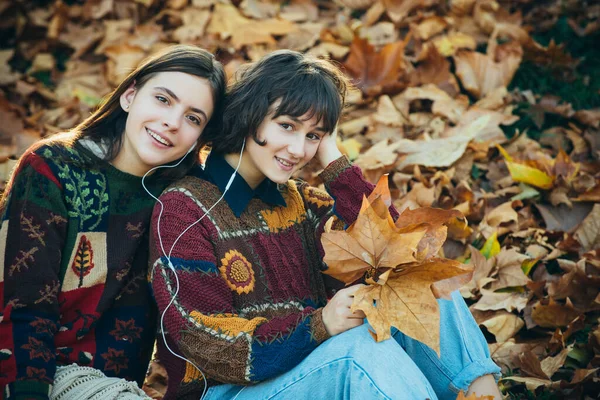 秋の葉の上に座って幸せな女の子。秋の屋外活動。秋の葉の花束を持つ2人の女の子。秋の公園で2人の美しい女の子の友人。秋の公園で二人の陽気な女性. — ストック写真