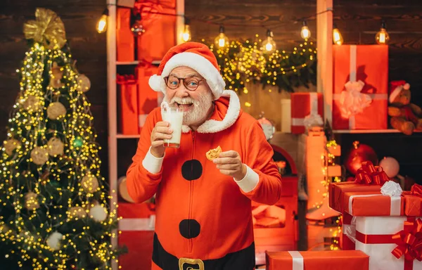 Weihnachtsmann mit Bart und Schnurrbart. Der Weihnachtsmann macht lustige Gesichter und pflückt Plätzchen. Porträt des Weihnachtsmannes mit Schokoladenkeks und einem Glas Milch. — Stockfoto