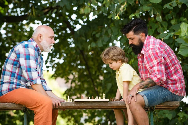 Babası ve dedesi olan bir çocuk. Küçük çocuk büyükbabası ve babasıyla satranç oynuyor. Yaz ve aktif tatiller. Son sınıf öğrencisi satranç oyunundaki bir sonraki hamlesini düşünüyor.. — Stok fotoğraf