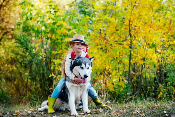 Dziewczyna w kapeluszu przytula psa. Mała dziewczynka ze swoim psem, Alaskan Malamute, pełna długość. Dziecko i pies na tle natury. Beztroskie dzieciństwo. Natura spaceruje z dziećmi i zwierzątkiem. Dzieci z uroczym psem. — Zdjęcie stockowe