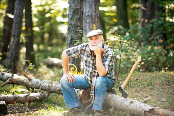 Old bearded man in forest. Fashion portrait man. Aged funny forester. Woodman in forest. Old bearded man with axe. Bearded male in hat on a background of trees. Mature farmer with beard in hat. — Stock Photo, Image