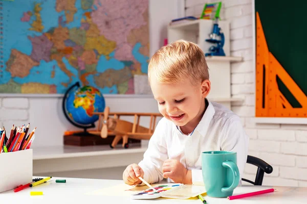 子供は屋内の机に座っている。面白い顔の学校の仕事を持つかわいい生徒。子供は学校に行く準備ができて. — ストック写真