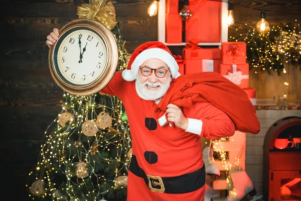 Der lächelnde Weihnachtsmann zeigt den Wecker mit fünf Minuten vor zwölf. Weihnachtsmann - bärtiger, lustiger Senior. Porträt des überraschten und lustigen Weihnachtsmannes. Frohe Weihnachten. — Stockfoto