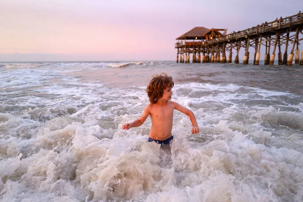 Feliz niño jugando en el mar al atardecer. Niño jugando al aire libre saltando al agua en las vacaciones de verano en la isla de playa tropical . — Foto de Stock