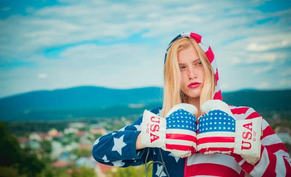 Deportiva patriótica con sudadera con capucha y guantes de boxeador con patrón de bandera americana en el fondo de la ciudad. Linda mujer rubia fuerte celebrando el Día de la Independencia de los EE.UU. . — Foto de Stock