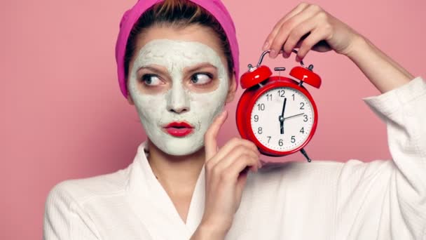 Funny girl with mask on face holds alarm clock in hand. Beautiful girl with cosmetic mask on face. Woman wearing face mask. Face care concept. — Stock Video