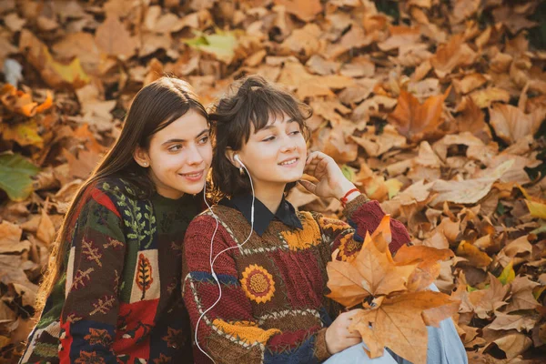 Krásné dívky sedí ve žlutém listí v podzimním parku. Dvě usměvavé ženy v podzimním parku. Dvě holky se žlutým listím. Portrét dvou roztomilých dívek sedících na listí v podzimním parku. — Stock fotografie