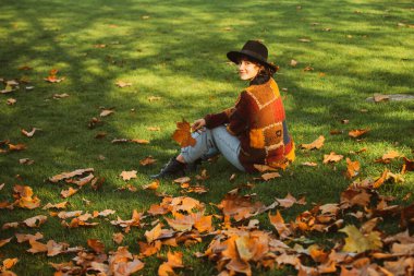 Sarı akçaağaç yaprağı tutan bir kadın. Moda insanları. Canlı yeşilliklerle çevrili ilham verici bir kız. Yaprak buketi tutan hayalperest kız. Arka plandaki kadın ağaç yapraklarından dökülüyor.