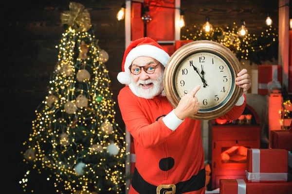 Weihnachtsmann - bärtiger, lustiger Senior. Fröhlicher Weihnachtsmann mit Wecker am Kamin und Weihnachtsbaumhintergrund. Lächelnder Weihnachtsmann zeigt Wecker mit fünf Minuten vor zwölf. — Stockfoto