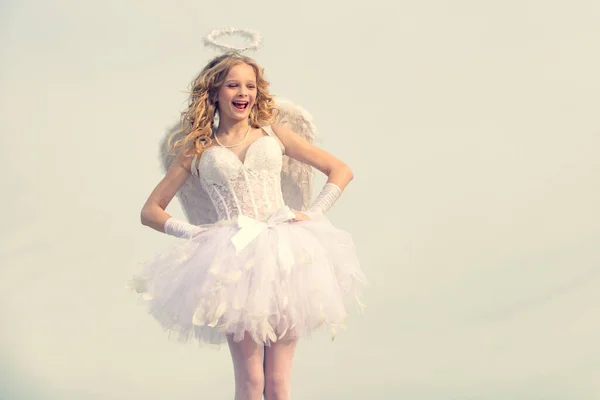 Chica inocente con alas de ángel de pie con arco y flecha contra el cielo azul y nubes blancas. Retrato de una niña cupido rezar. Niño con carácter angelical . —  Fotos de Stock