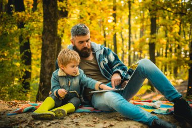 Evde eğitim doğa konsepti. Ebeveynler bebeklere dizüstü bilgisayar kullanmayı öğretiyor. Sonbahar parkında dışarıda vakit geçiren mutlu baba ve oğul. Çocuk ve babası sonbahar parkında. Hem baba hem de çocuk...