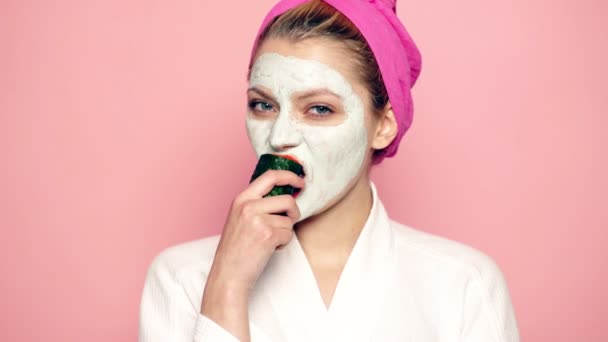 Chica con una máscara en su cara comiendo pepino sobre fondo rosa. Mujer con mascarilla. Cuidado facial, concepto de belleza facial . — Vídeos de Stock