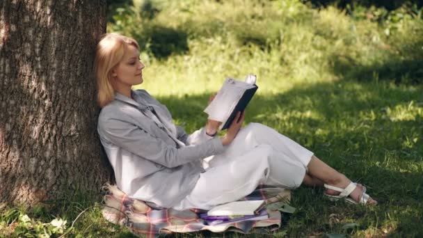 Το κορίτσι διαβάζει από το βιβλίο, κάθεται στο γρασίδι στο πάρκο. Μια μαθήτρια ετοιμάζεται για εξετάσεις στο πάρκο. Η έννοια της υπαίθριας εκπαίδευσης. — Αρχείο Βίντεο