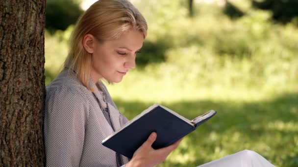 Flickan läser ur en anteckningsbok som sitter på gräset i parken. Flickelev förbereder sig för lektioner i parken. Begreppet utomhusutbildning. — Stockvideo