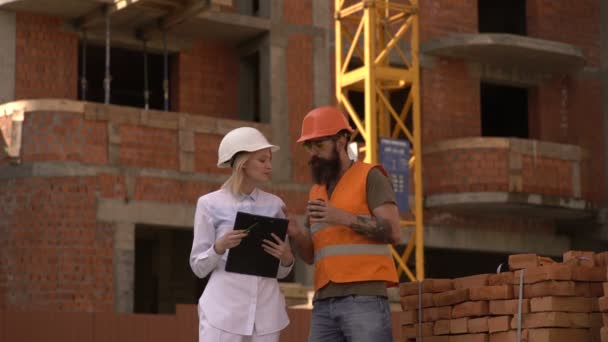 Due ingegneri stanno discutendo un piano di costruzione sul cantiere. Una donna col casco bianco e un uomo barbuto parlano sul cantiere. Concetto di costruzione. — Video Stock