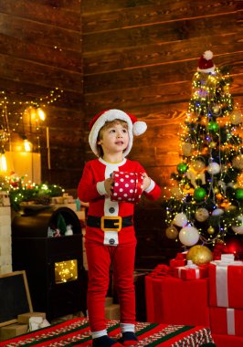 Mutlu çocuk Noel ağacı süslemesi. Noel ağacının yanında şirin bir çocuk. Noel süsleri. Çocuklar tatilin tadını çıkarır. Noel çocuğu elinde kırmızı bir hediye kutusu tutuyor.