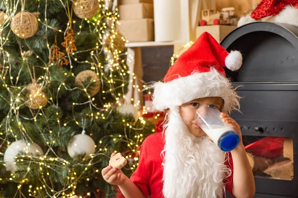 Mikołaj trzyma ciasteczko i szklankę mleka na tle choinki. Mleko i pierniki ciasteczko dla Mikołaja przed Christmas light bokeh tle. — Zdjęcie stockowe