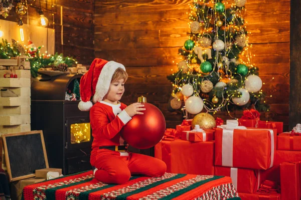 Glückliches Kind trägt Nikolauskleidung und schmückt die Christbaumkugel. Weihnachtskonzept. Weihnachtsmann hilft Kindern. — Stockfoto