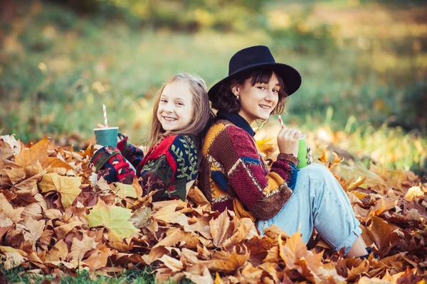 Irmãs felizes com folhas amarelas de outono. Folhas amarelas caídas. Rapariga sorridente com irmã com uma folha de laranja nas mãos sentada na folhagem de outono na floresta. Irmãs desfrutando natureza queda . — Fotografia de Stock