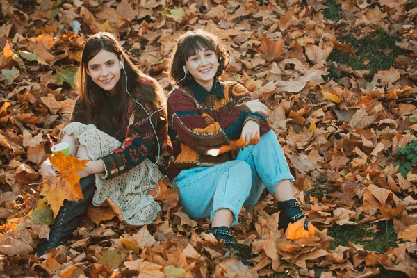 Dos chicas sonriendo en el parque de otoño. Lindos jóvenes estudiantes se sientan en las hojas de otoño. Chica con su mejor amiga. Dos chicas en un parque. Retrato de dos mujeres riendo al aire libre en el parque de otoño . — Foto de Stock