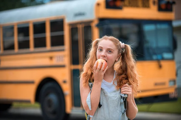 Bildung: Lächelndes Studentenmädchen bereit für den Bus Schulmädchen vor dem Schulbus beim Apfelessen. — Stockfoto