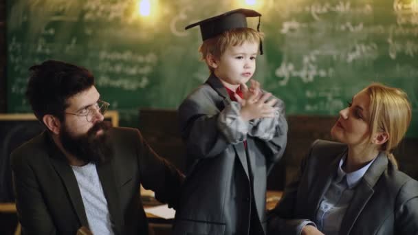 Divertente ragazzino in berretto di laurea si trova vicino a un consiglio scolastico con gli insegnanti. Bambino a scuola. All'asilo con i genitori. Insegnanti e alunni in classe. Carino bambino che studia con padre e madre. — Video Stock