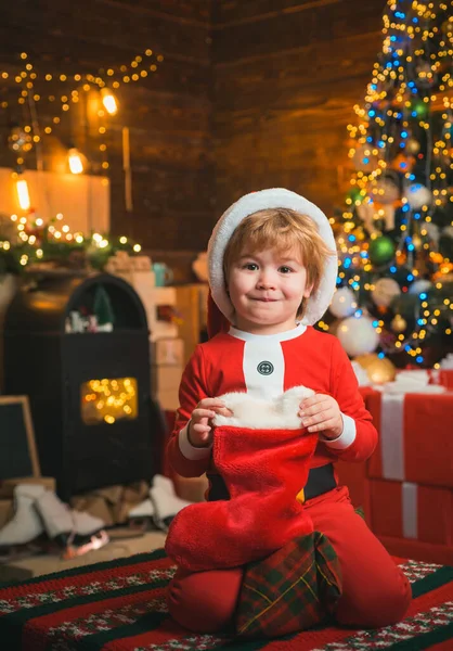 木製の背景にクリスマスソックスを着た子供。幸せな小さな笑顔の少年サンタ服を着ている。幸せな子供は贈り物を楽しんでいます。クリスマスツリーの背景. — ストック写真