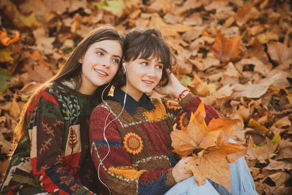 Dos chicas divirtiéndose en el parque. Hermosas chicas jóvenes con el pelo largo con un ramo de hojas amarillas en las manos en el parque de otoño. El territorio está completamente cubierto por el follaje caído. — Foto de Stock