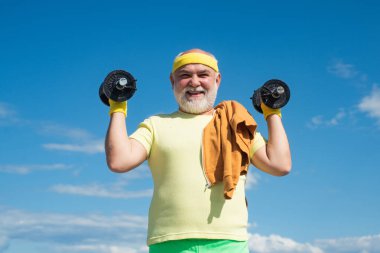 Kıdemli sporcu mavi gökyüzü arka planında dambıl kaldırarak egzersiz yapıyor. İzole edilmiş, kopyalama alanı.