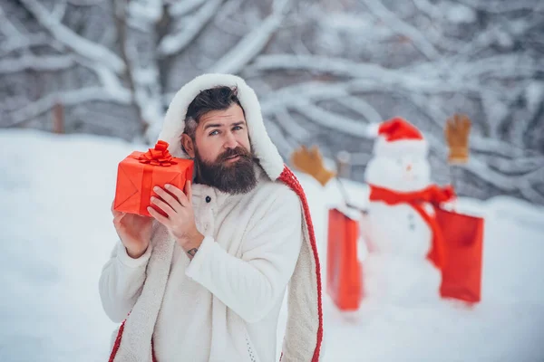 Retrato de inverno de hipster bonito na neve Jardim com presente fazer boneco de neve. Homem de Natal e boneco de neve no fundo branco da neve. Papai Noel bêbado em casa . — Fotografia de Stock
