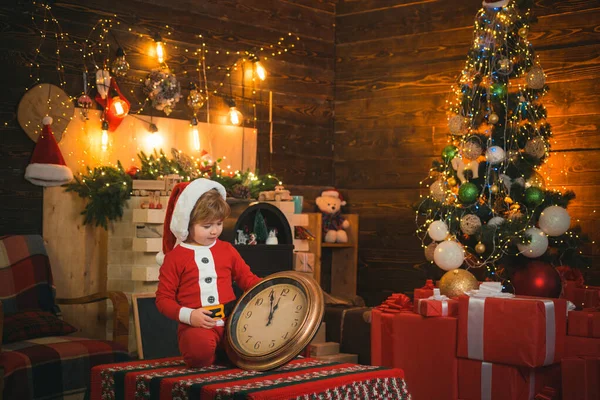 Sonriente niño ayudante de Santa mira el reloj y la espera de Año Nuevo medianoche en el fondo decorado de Navidad de lujo. Feliz Navidad y feliz año nuevo. Interior brillante Año Nuevo. — Foto de Stock