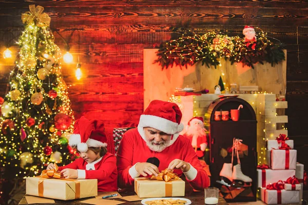 プレゼント付きのクリスマスプレゼントを保持サンタクロースとサンタのヘルパー。メリークリスマスと幸せな新年。小さなサンタクロースはクリスマスの背景に贈り物を開きます. — ストック写真
