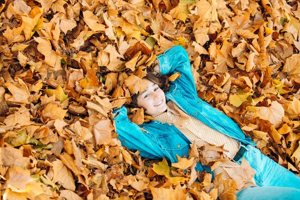 Mulher de outono deitada sobre folhas e sorrindo. Jovem com folhas de outono na mão e queda de fundo de jardim de bordo amarelo. Menina sorridente coleta folhas amarelas no outono . — Fotografia de Stock