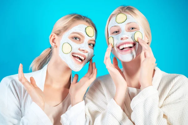 Jonge gelukkige meisjes doen gezichtsmasker met plakjes komkommer op blauwe achtergrond. Huidverzorging en -behandeling, spa, natuurschoon en kosmetologie. — Stockfoto