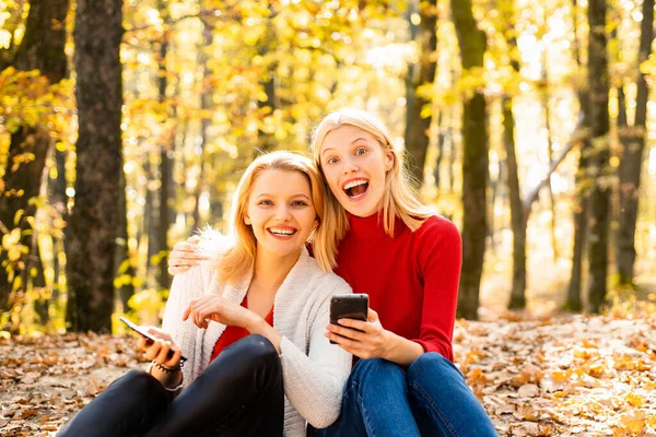 Jovens meninas felizes segurando telefone celular e sentado em folhas caídas ao ar livre. Mulheres jovens no Outono. Estudantes amigos no belo tempo de outono na natureza . — Fotografia de Stock