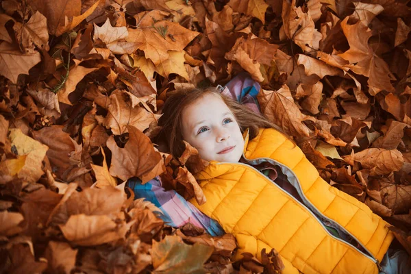 Criança feliz deitada em folhas de outono. Um miúdo a brincar no parque de Outono. Menina feliz adorável com as folhas caídas. A posar. Miúdo deitado em folhas. Brincadeira de criança com folhagem de bordo dourada . — Fotografia de Stock
