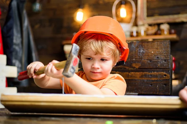 Kid hammers nails with a hammer in a wooden board. Boy child busy in helmet learning to use hammer. Child hammering nail into wood at home.
