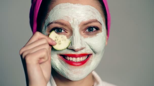 Sluit een jonge vrouw met een gezichtscrème op haar gezicht. Grappig meisje, huidverzorging. Model met een gezichtsmasker en een handdoek op haar hoofd die haar ogen sluit met komkommers. — Stockvideo