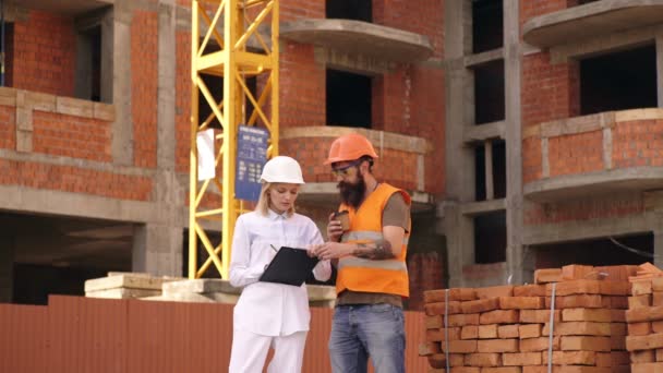 Frau mit Helm und Ordner kommuniziert vor dem Hintergrund der Bauarbeiten mit einem bärtigen Mann in Schutzweste. Ingenieur kommuniziert mit dem Auftragnehmer auf der Baustelle. — Stockvideo