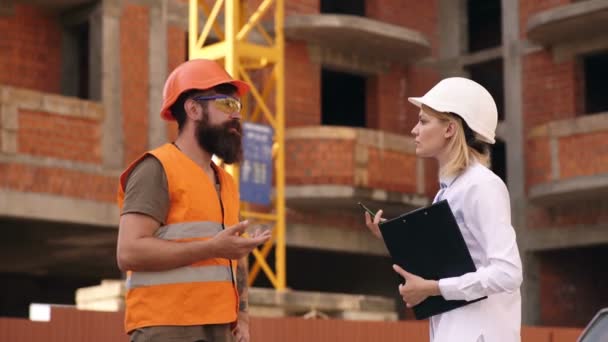 Un homme barbu dans un casque et un gilet de protection communique avec une femme dans un casque et un dossier dans ses mains sur le chantier de construction. Concept de construction. — Video
