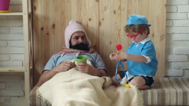 Um rapazinho com o pai no hospital. Médico de família. Medicina e saúde. Pai e filho de uniforme médico brincam de médico paciente. Pequeno médico trata seu paciente, que está deitado debaixo de um cobertor, com pílulas . — Vídeo de Stock