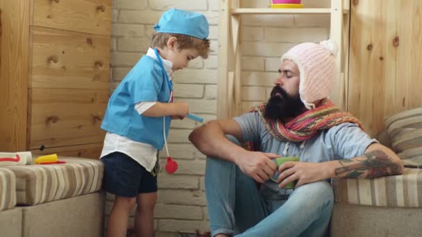 Father and son play the doctor patient. Little doctor with stethoscope examining sick bearded man. — Stock Video